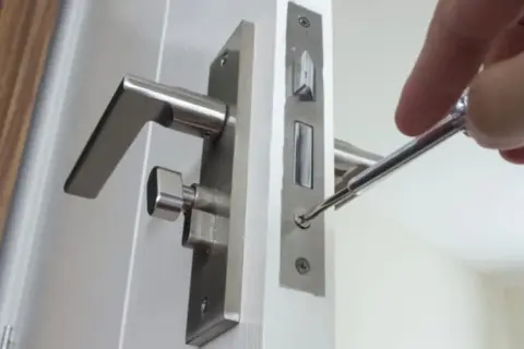 Réparation d'une poignée de porte qui claque avec le vent. Un tournevis ajuste la serrure.