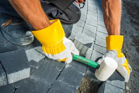 Pracownik w żółtych rękawicach układa kostkę brukową na betonie, używając gumowego młotka.