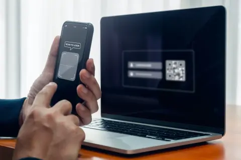 Ręce trzymają telefon z napisem "SCAN TO LOGIN", obok laptop z kodem QR. Szybkie logowanie, jak zeskanować kod na telefon.