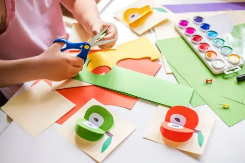 Manos pequeñas cortando papel para manualidades para niños de 3 a 5 años. Pinturas, tijeras y frutas de papel decoran la mesa.