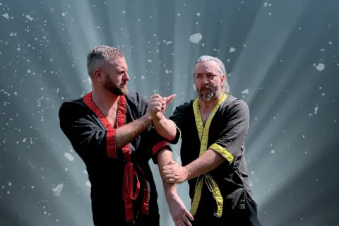 Två män i traditionella dräkter tränar wing chun. Ljuset strålar bakom dem.