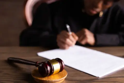 Młotek sędziowski i dokumenty na biurku. Referendarz sądowy pochyla się nad papierami, pisząc.