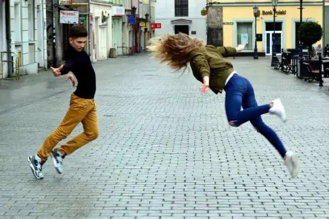 Młoda para tańczy na ulicy, ona unosi się w powietrzu, on biegnie. Ich ruchy przypominają taniec belgijka.