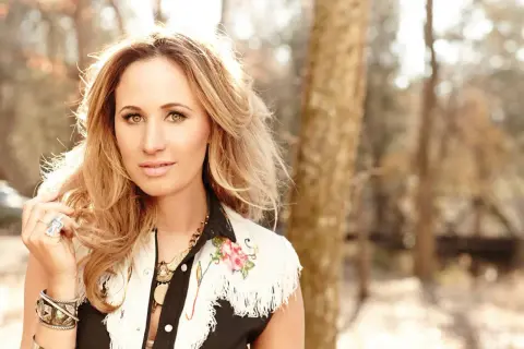 Jessi Alexander, a blonde woman with striking eyes, poses outdoors in a fringed vest adorned with floral embroidery.