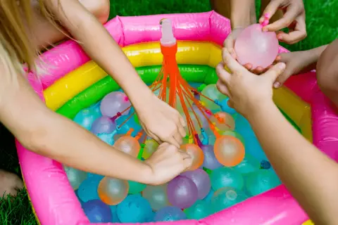 Manos infantiles llenando globos de agua en una piscina inflable multicolor. ¡Diversión garantizada con estos juegos de agua!