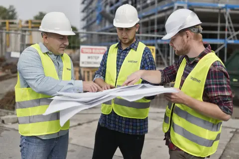 Trzech budowlańców w kaskach i kamizelkach omawia plany. Jeden z nich wskazuje palcem na dokumenty, ustalając szczegóły projektu i prowizję.