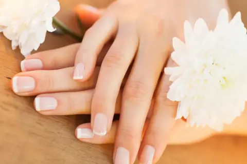 Delikatne dłonie z francuskim manicure, otoczone białymi kwiatami. Paznokcie wyglądają zdrowo, nie są suche.