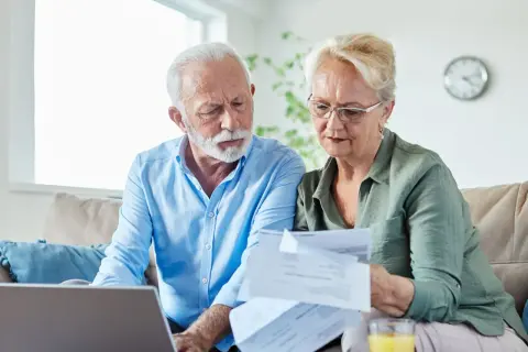 Couple âgé examinant des papiers, peut-être pour une aide EHPAD GIR 2. Ils sont sur un canapé avec un ordinateur portable.