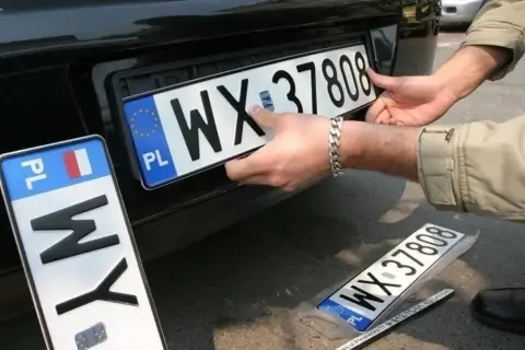 Montaż nowych tablic rejestracyjnych. Jedna z nich, z numerem WX 37808, jest właśnie wkładana. Obok leży kolejna, zapakowana.