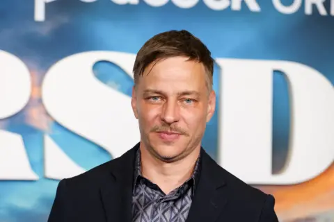 Tom Wlaschiha, mit einem Schnurrbart und blauen Augen, posiert vor einem Hintergrund mit großen weißen Buchstaben. Er denkt vielleicht an seine tom wlaschiha kinder.