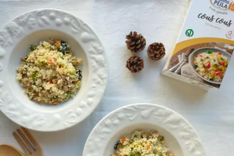Piatti di cous cous con verdure e ceci, perfetti per le ricette cous cous invernale. Accanto, una confezione di cous cous e pigne decorative.