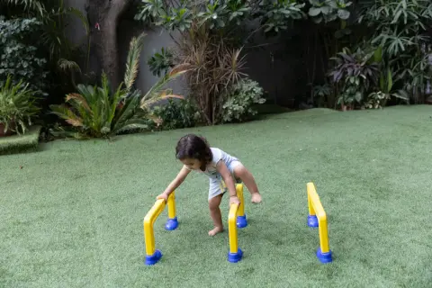A toddler practices gross motor outdoor activities, balancing on yellow and blue hurdles in a grassy yard.