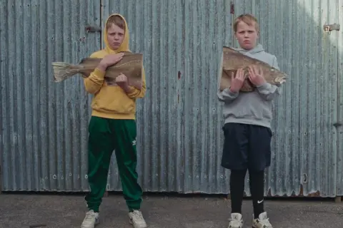 Två barn, som påminner om Sverrir Gudnason som barn, håller varsin fisk framför en plåtvägg.