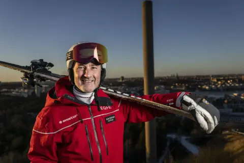 Ski-entusiast med skidor på axeln, klädd i röd jacka med SkiStar-logga. Han ser ut att vara en del av SkiStar ledning, redo för äventyr.