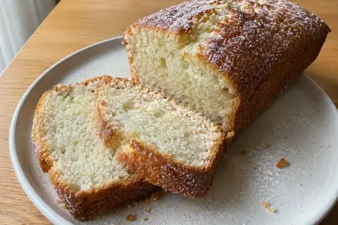 En saftig vegansk sockerkaka, pudrad med florsocker, ligger på en tallrik. Två skivor är upplagda bredvid.