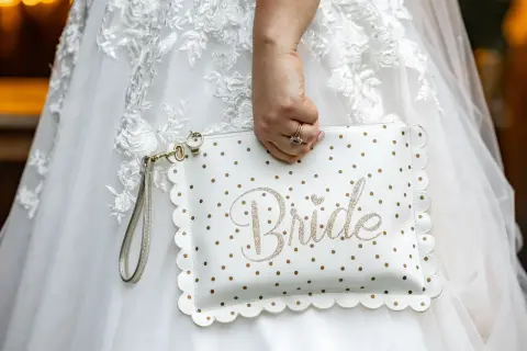 Biała kopertówka w złote kropki z napisem "Bride" to niezbędnik panny młodej. Dopełnia suknię ślubną.
