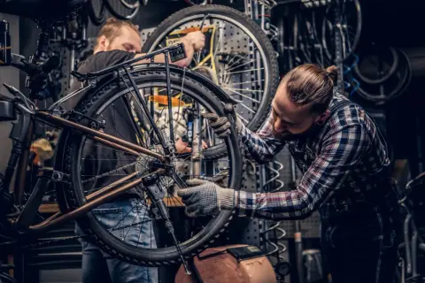 Dwóch mechaników pracuje nad rowerem w warsztacie. Jeden z nich dokonuje konserwacji roweru, dokręcając śruby kluczem.