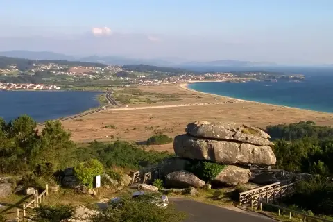 Vista panorámica desde el mirador de Siradella. Rocas gigantescas y un puente de madera en primer plano, con un paisaje de costa, mar azul y un pueblo al fondo.
