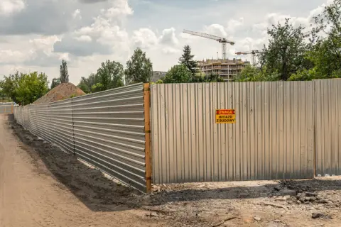 Metalowe ogrodzenie budowy, spełniające wymagania bezpieczeństwa, z tabliczką "Uwaga, wyjazd z budowy". W tle budynki i dźwigi.
