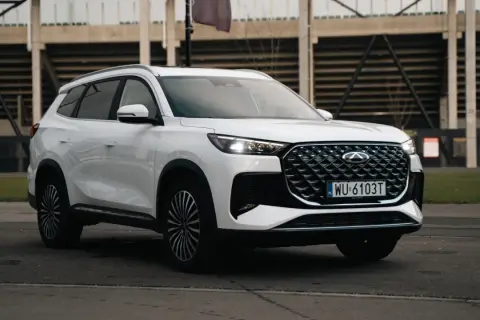 Biały Chery Tiggo 8 z charakterystycznym grillem i światłami LED, zaparkowany przed trybunami stadionu.