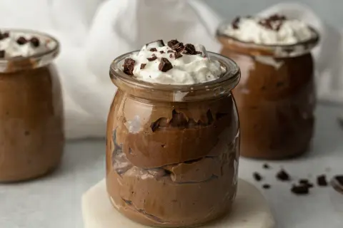Czekoladowy pudding proteinowy w słoiku, udekorowany bitą śmietaną i kawałkami czekolady.