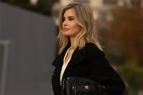Kobieta z beżowym blondem, w czarnym płaszczu i z pikowaną torebką Chanel, prezentuje elegancką stylizację.