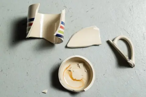 Rozbite fragmenty ceramicznego kubka z kolorowymi paskami leżą na szarym tle. Widać podstawę, korpus, uchwyt i kilka mniejszych kawałków. To materiał do nauki, jak skleić kubek ceramiczny.