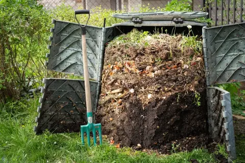 Ein grüner Komposter ist geöffnet und zeigt eine Füllung aus Gartenabfällen. Eine Mistgabel steht daneben, bereit, beim Kompost bauen zu helfen.