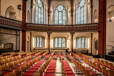 Innenraum einer epo church mit vielen roten Stuhlreihen und großen Bogenfenstern. Ein Mann sitzt allein.