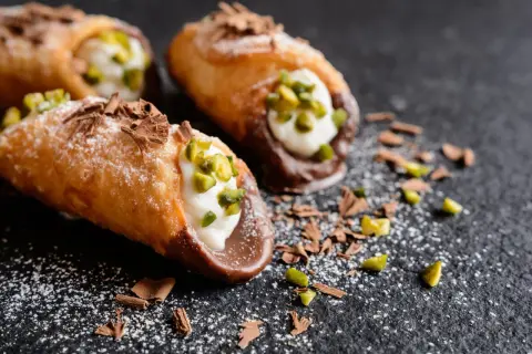 Trois cannoli salés, garniture crémeuse, pistaches concassées et copeaux de chocolat, saupoudrés de sucre glace sur fond sombre.