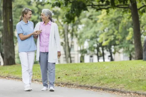 Pflegekraft stützt ältere Dame, die wegen Demenz weglauftendenz hat. Hilfsmittel wie diese Spaziergänge sind wichtig.