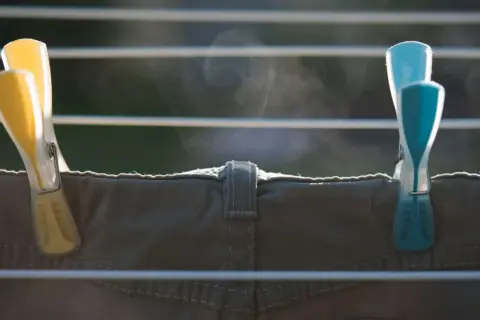 Zwei Wäscheklammern, gelb und blau, halten eine Hose auf einer Wäscheleine. Dampf steigt auf, die Hose trocknet schnell.
