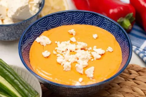 Cremiger Paprika-Feta-Dip in blauer Schüssel, garniert mit Feta-Käse. Dazu Gurkensticks und rote Paprika.
