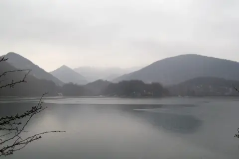 Paisaje brumoso con montañas cubiertas de árboles y el **embalse de Lareo** en primer plano. Ramas desnudas enmarcan la serena vista.