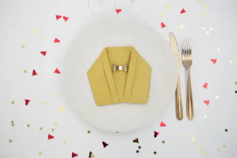 Pliage serviette nouvel an en forme de veste, orné d'un nœud papillon doré, sur une assiette blanche. Confettis sapins et étoiles rouges et dorés.