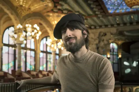 David Ucles sonríe con un gorro y una camiseta a rayas, en un salón con vidrieras y lámparas doradas.