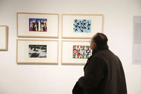 Un hombre observa obras de arte en una galería, incluyendo una que representa figuras militares y banderas, evocando el estilo de Pablo Serrano.