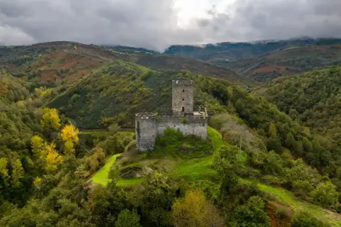 El **castillo de Doiras** se alza majestuoso sobre una colina verde, rodeado de montañas boscosas y un cielo nublado.