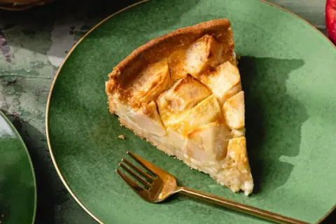 Une part de tarte normande aux pommes, dorée et appétissante, sur une assiette verte avec une fourchette dorée.