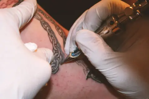 Manos enguantadas tatuando una cadena rota en la piel.