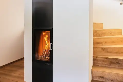 Ein moderner Kamin mit brennendem Feuer, daneben eine Holztreppe. Hier sieht man, was ein Grundofen leisten kann: Wärme und Gemütlichkeit.