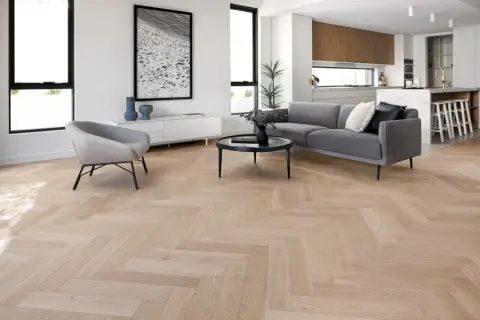 Elegante salón con suelo de madera en espiga, sofá gris, sillón y cocina moderna al fondo. La colocación espiga resalta la calidez del espacio.