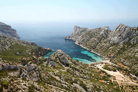 Vue aérienne de la calanque de Sormiou, un joyau bleu turquoise niché entre des falaises rocheuses. L'itinéraire pour y accéder est visible, serpentant à travers la végétation méditerranéenne.