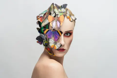 Portrait d'une femme au maquillage audacieux, ornée de papillons colorés sur la tête et le visage, évoquant une **flora borsi** onirique.