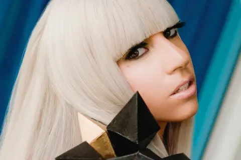 Lady Gaga, con capelli biondo platino e un look audace, cattura l'essenza del suo **inizio carriera** con un'espressione intensa e un accessorio geometrico.