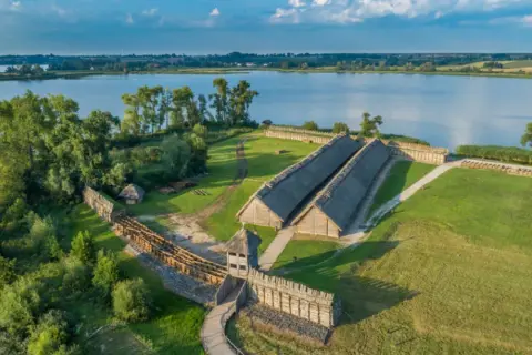 Rekonstrukcja osady Biskupin z drewnianymi domami krytymi strzechą, wałami obronnymi i jeziorem w tle.