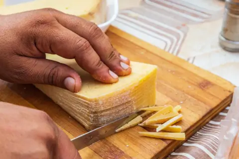 Mano cortando un bloque de queso en finas láminas. La corteza dorada se ve apetitosa, demostrando que se puede comer la corteza del queso.