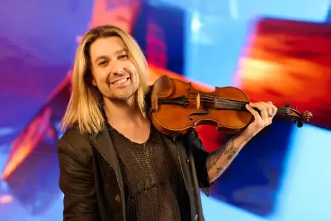 Ein lächelnder Mann mit langen blonden Haaren hält eine Geige. Er könnte der beste Geiger der Welt sein, mit seiner Leidenschaft für Musik.