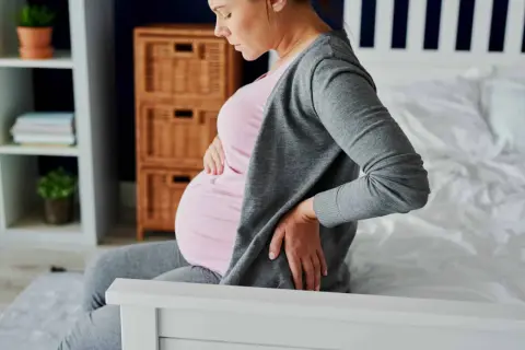 Femme enceinte assise sur un lit, tenant son ventre et se tenant le bas du dos, souffrant peut-être d'hémorroïdes.