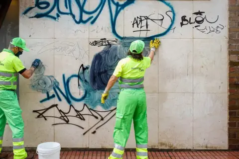 Dos trabajadores con uniformes verdes brillantes realizan la limpieza de grafitis en una pared de mármol.